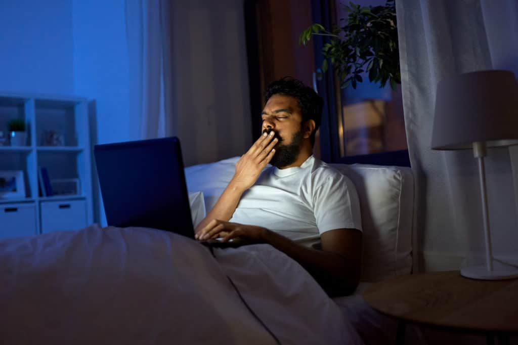 a man yawns while watching videos on a laptop in bed late at night