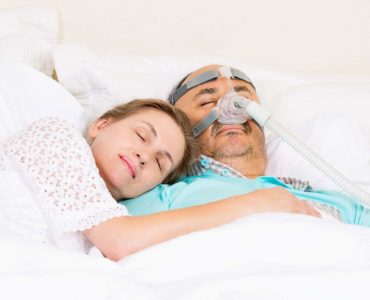 A couple sleeps in bed and the man is wearing a CPAP mask.